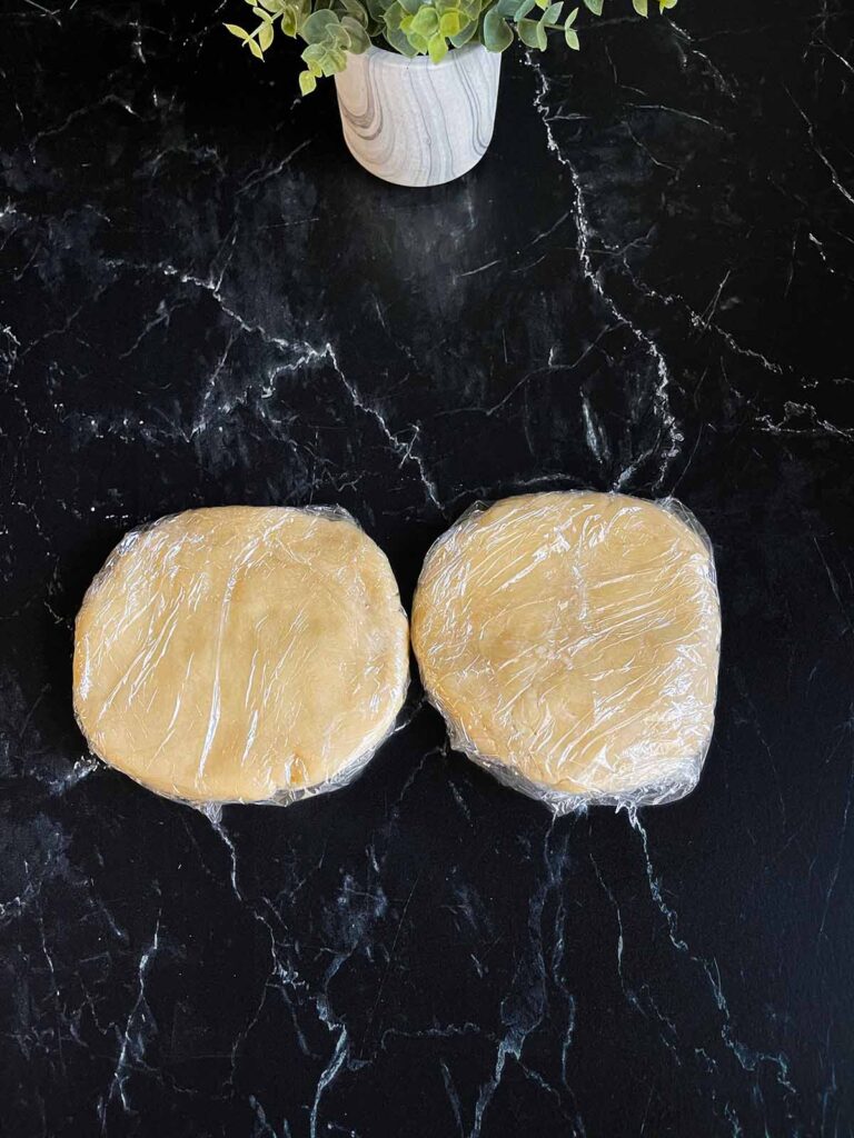 Pie crust dough wrapped in plastic wrap on a dark surface.