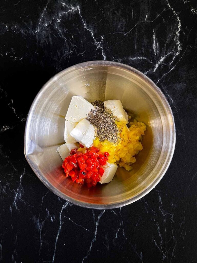 Ingredients for a cream cheese mixture for italian pinwheels in a silver mixing bowl on a dark surface.