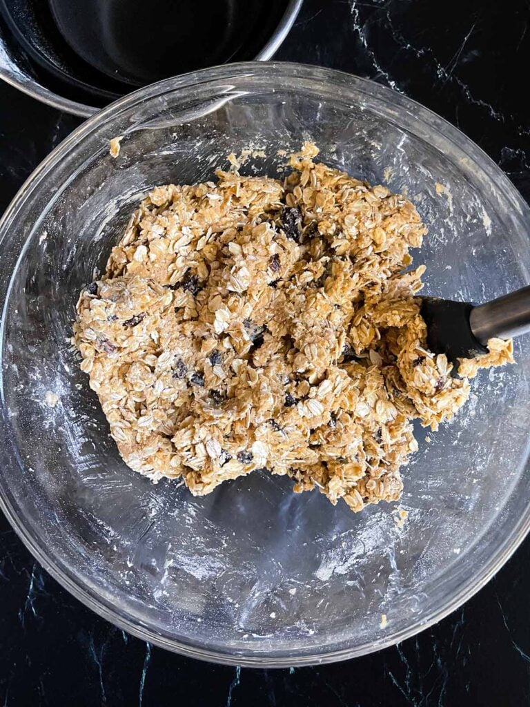 Old fashioned oats and raisins stirred into the cookie dough in a glass bowl on a dark surface.