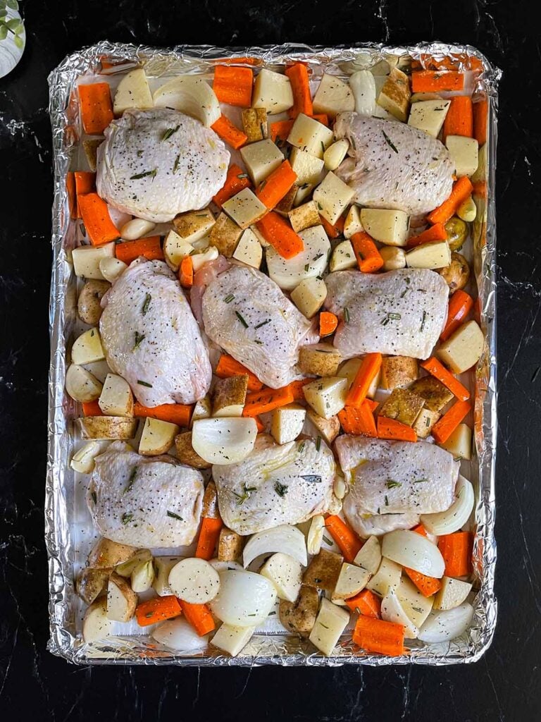 Seasoned raw chicken and vegetables in a foil and parchment paper lined rimmed baking sheet.