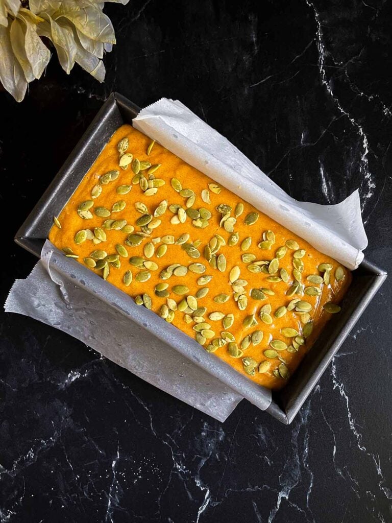 Pumpkin bread batter sprinkled with pumpkin seeds in a parchment paper lined loaf pan.