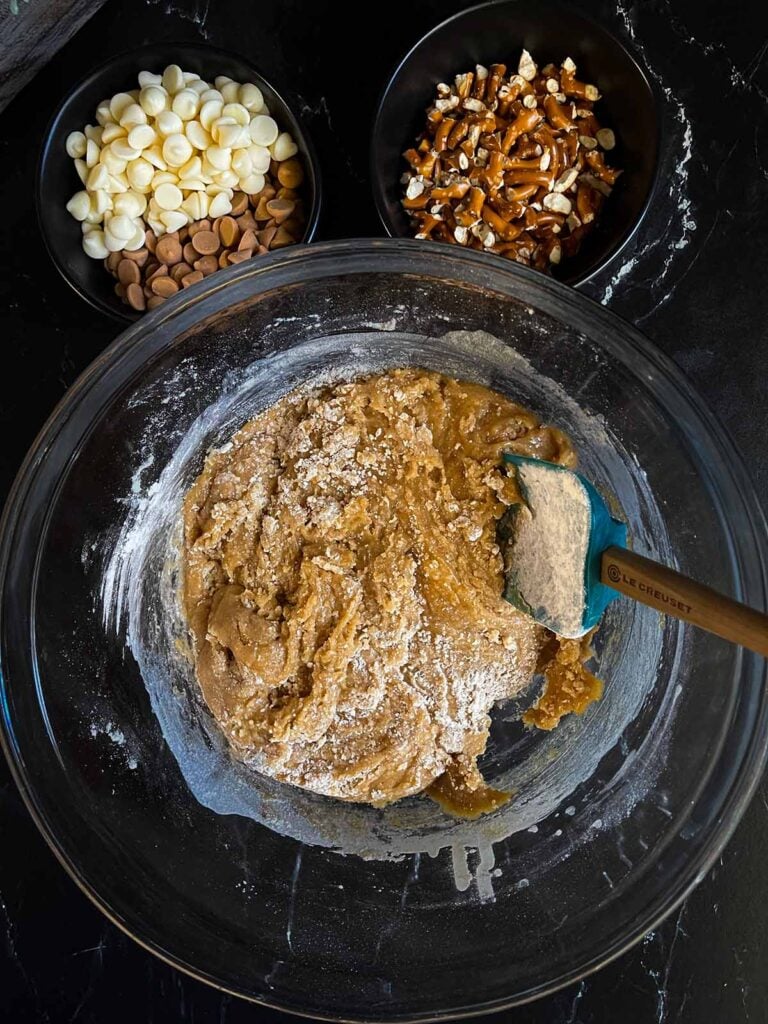 Mixing salted butterscotch pretzel cookie dough ingredients in a glass bowl on a dark surface.