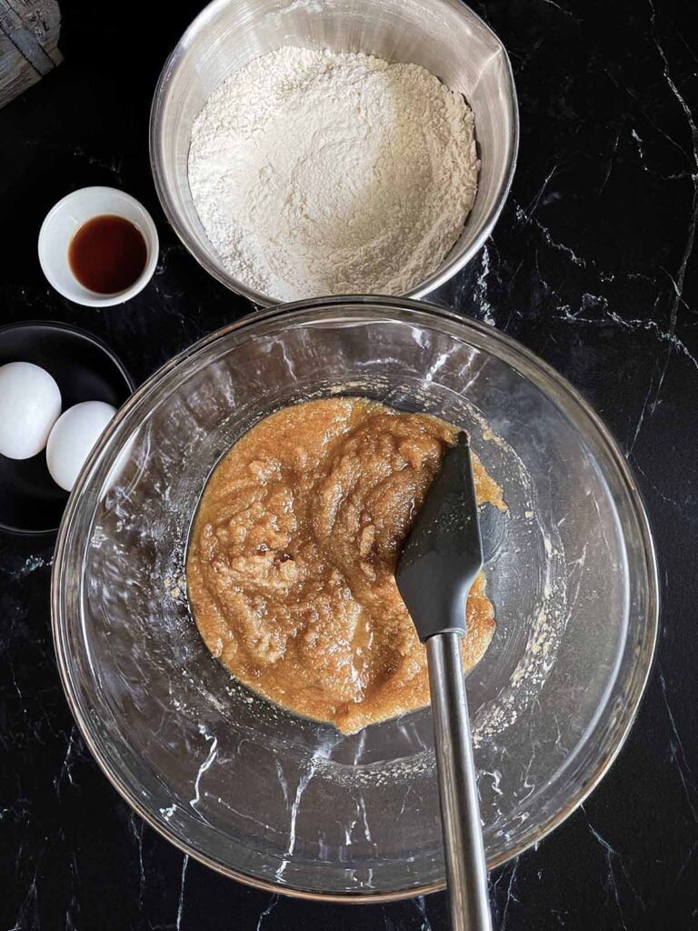 Wet ingredients for Snickerdoodle Bars in a large glass bowl with the dry ingredients in a smaller metal bowl on a dark surface.