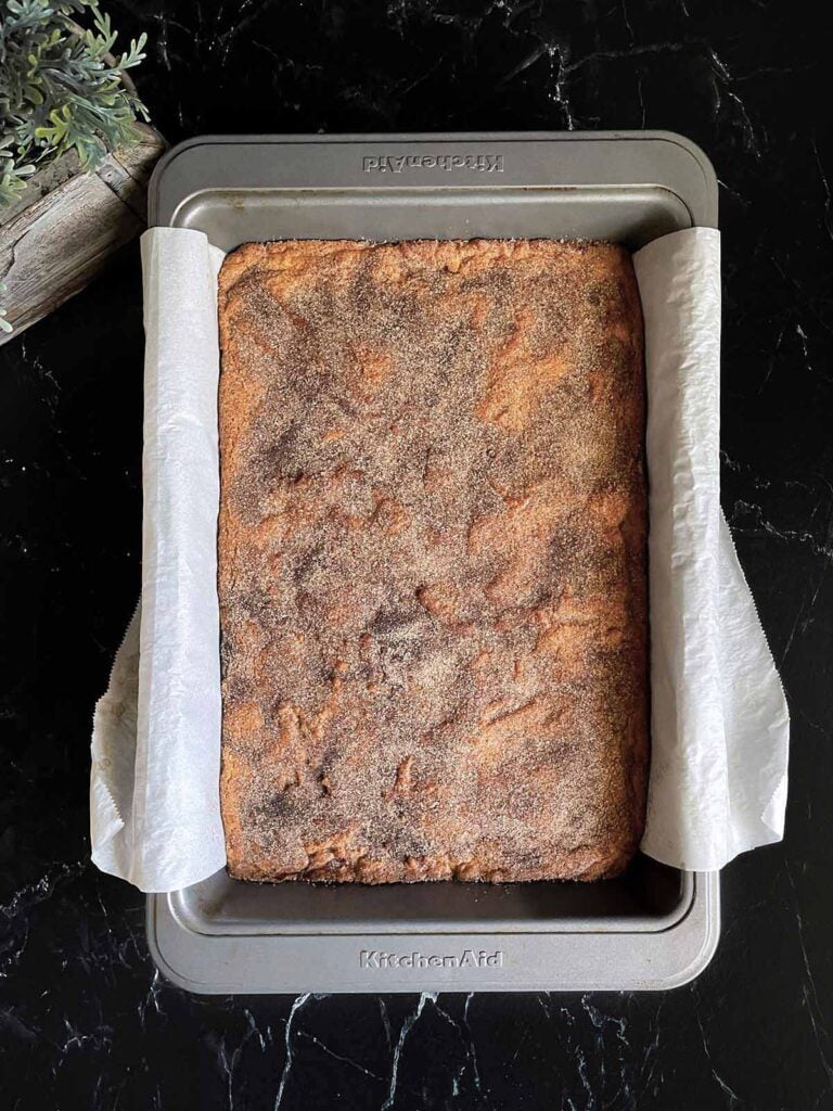 Baked Snickerdoodle Bars in the parchment paper lined baking pan on a dark surface.