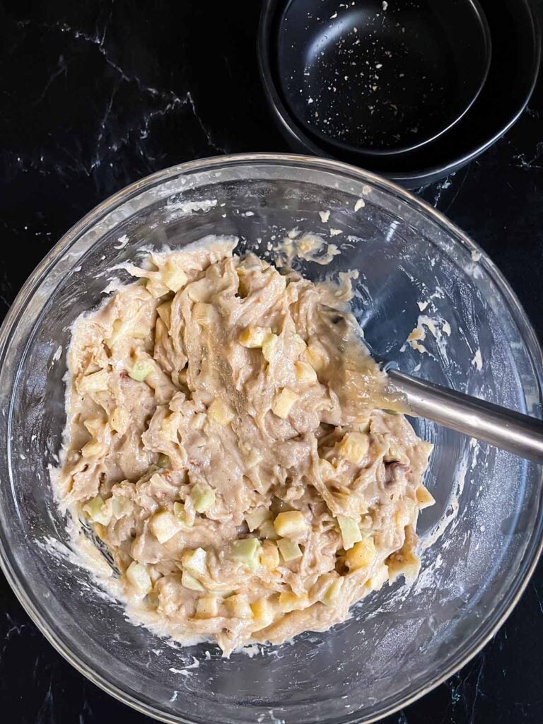 Apple Dapple Cake batter mixed in a glass mixing bowl on a dark suface.