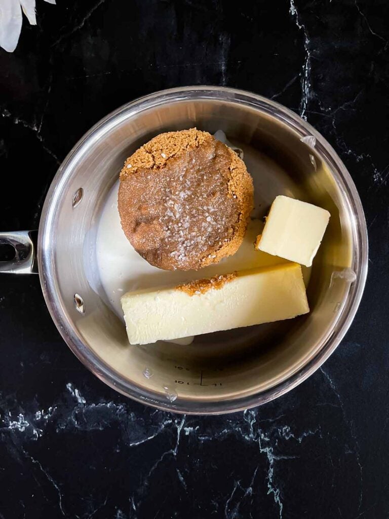 Brown sugar, butter, heavy cream, and kosher salt in a medium saucepan on a dark surface.