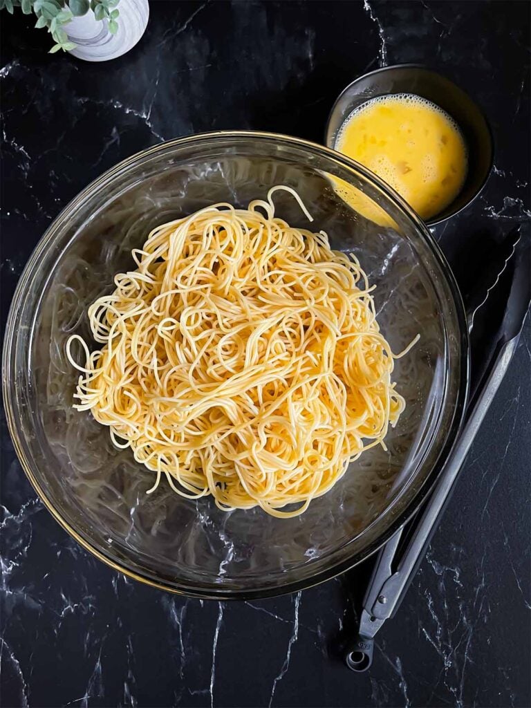 Boiled thin spaghetti in a large glass bowl with the beaten eggs in a small bowl on a dark surface.