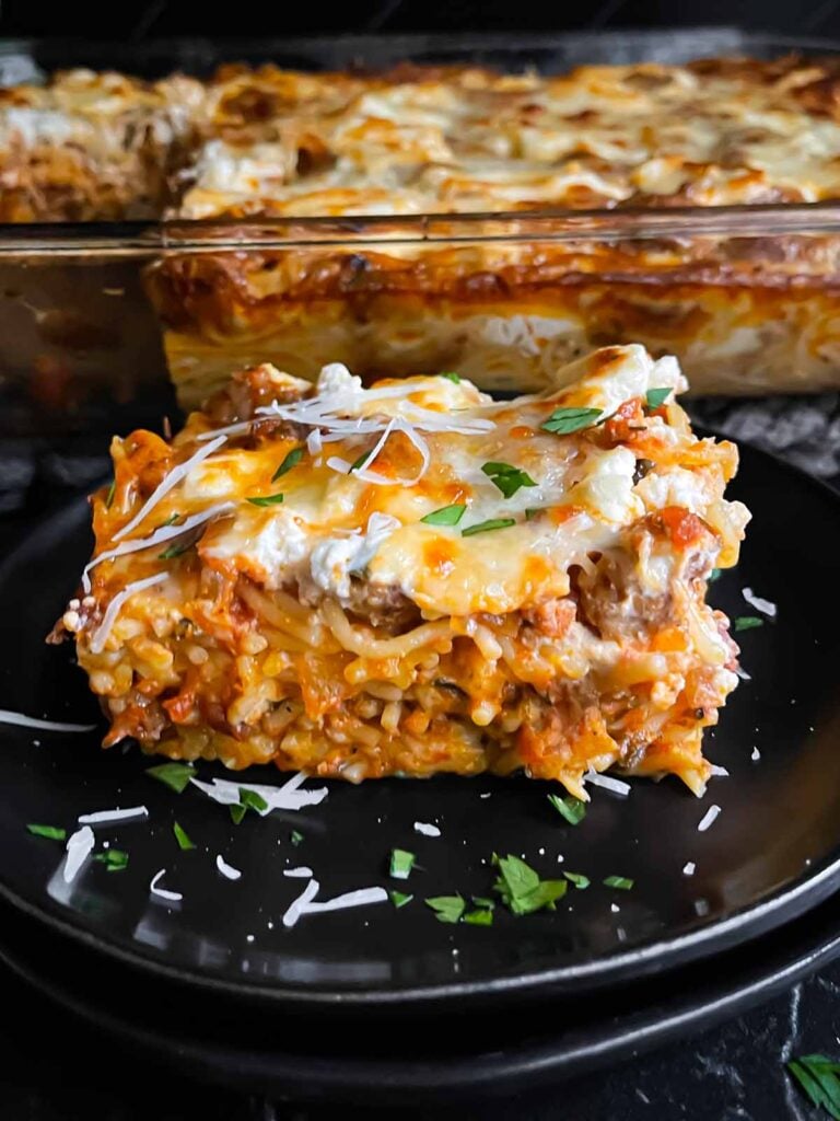 A serving of baked spaghetti garnished with parsley and Romano cheese on a dark plate on a dark surface.