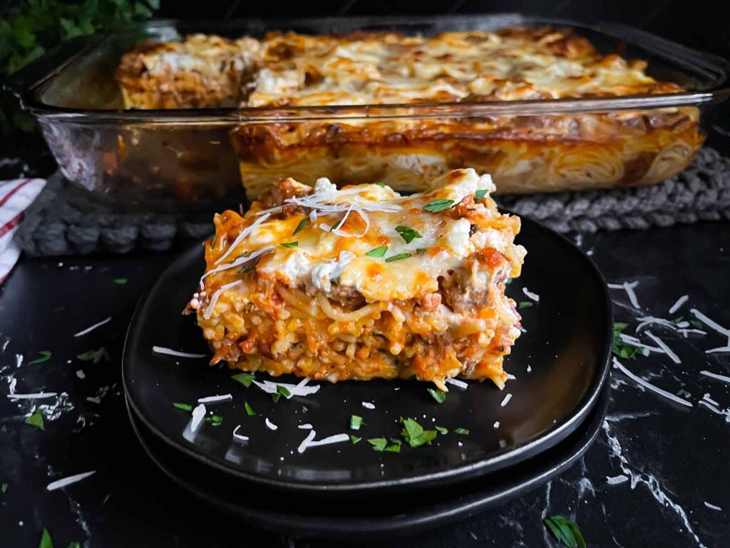 A serving of baked spaghetti garnished with parsley and Romano cheese on a dark plate on a dark surface.