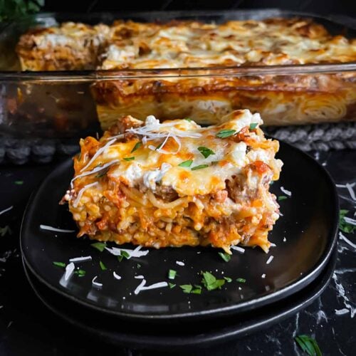 A serving of baked spaghetti garnished with parsley and Romano cheese on a dark plate on a dark surface.