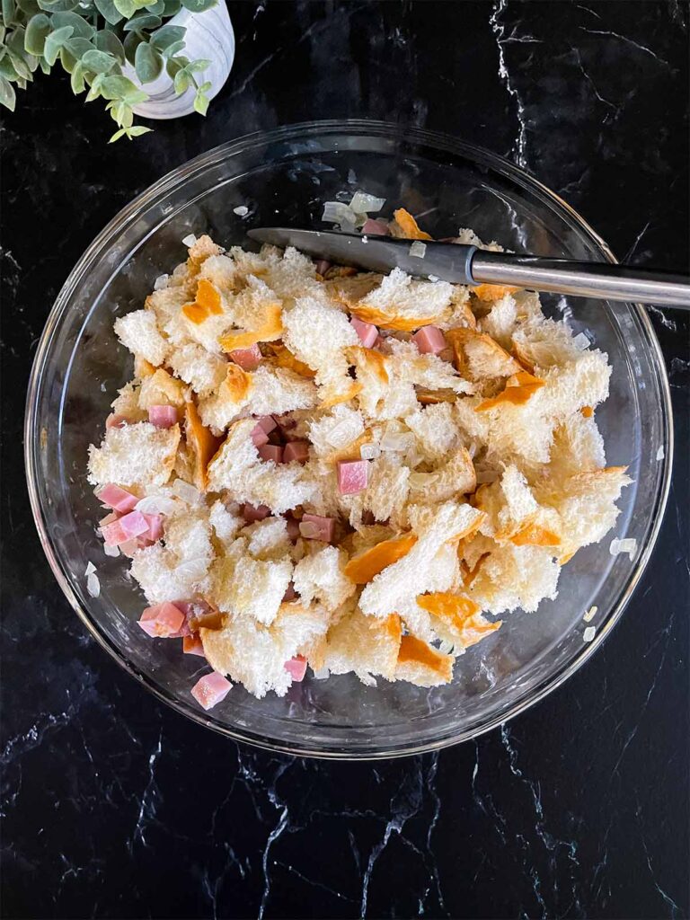 Torn bread pieces, onions, and ham in a large glass mixing bowl on a dark surface.