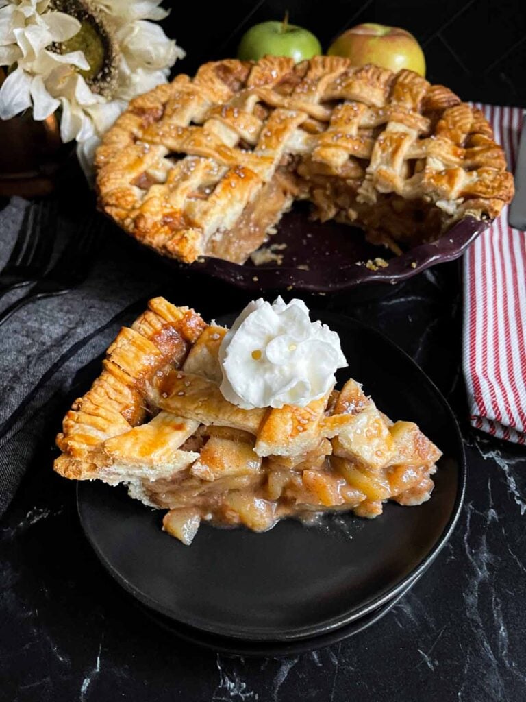 A slice of apple pie garnished with whipped cream on a dark plate on a dark surface.