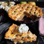 A slice of apple pie garnished with whipped cream on a dark plate on a dark surface.