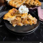 A slice of apple pie garnished with whipped cream on a dark plate on a dark surface.