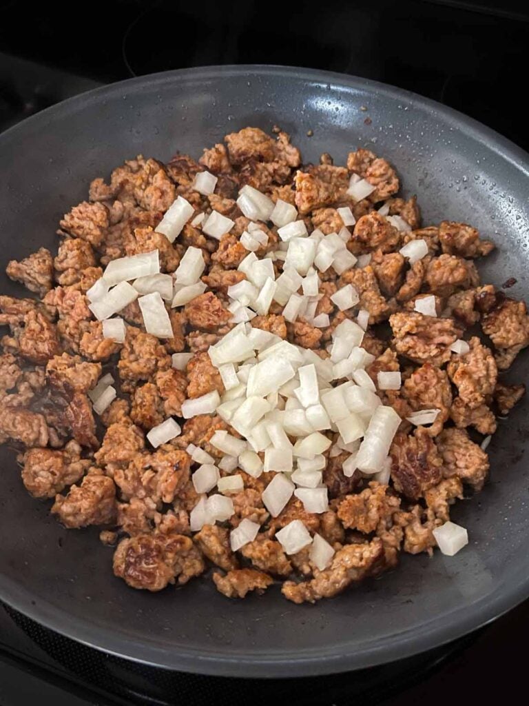 Onions added to browned italian sausage in a skillet.