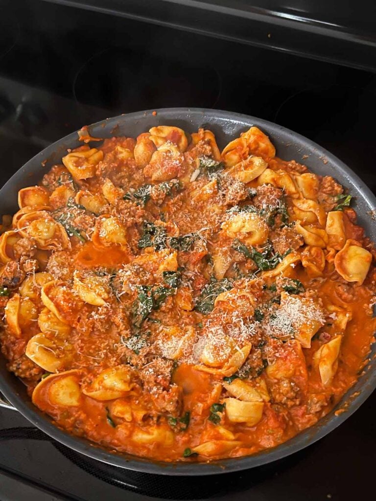 Adding cheese to tortellini with italian sausage and spinach in a skillet.