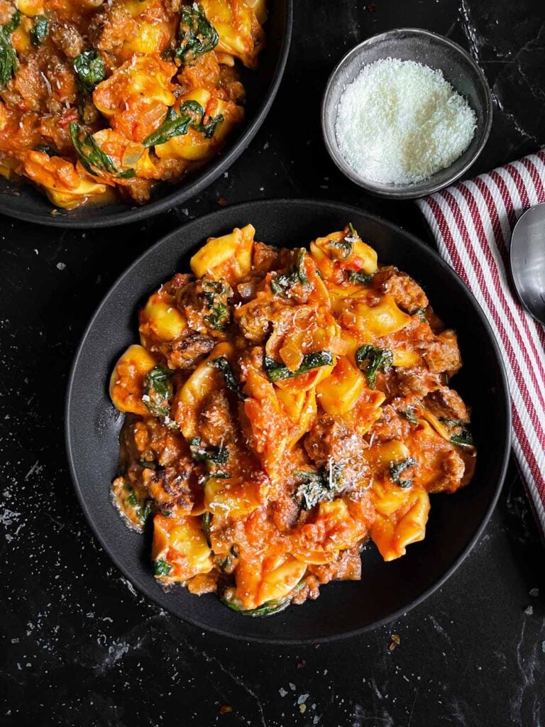 Cheese tortellini with sausage and spinach in a dark bowl on a dark surface.