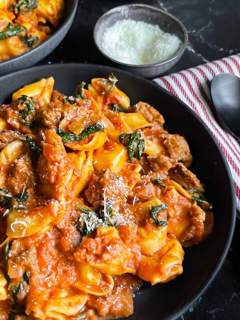 Tortellini with italian sausage and spinach in a dark bowl on a dark surface.