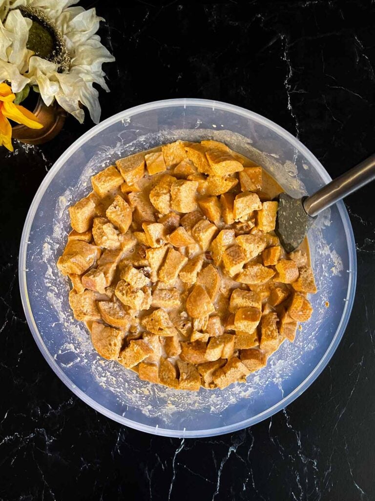 Bread mixed with the custard ingredients for pumpkin bread pudding in a large bowl on a dark surface.