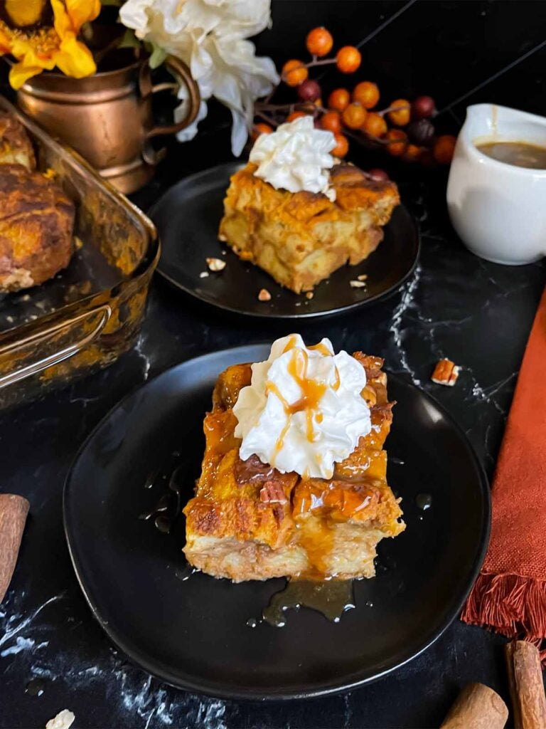 Slice of pumpkin bread pudding garnished with whipped cream and salted caramel sauce on a dark surface.