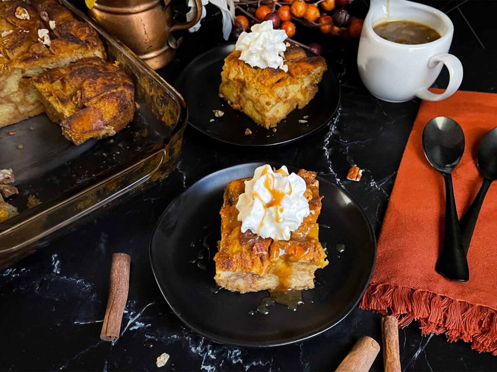 Slice of pumpkin bread pudding garnished with whipped cream and salted caramel sauce on a dark surface.