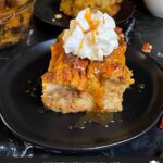 Slice of pumpkin bread pudding garnished with whipped cream and salted caramel sauce on a dark surface.