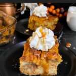 Slice of pumpkin bread pudding garnished with whipped cream and salted caramel sauce on a dark surface.