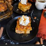 Slice of pumpkin bread pudding garnished with whipped cream and salted caramel sauce on a dark surface.