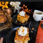 Slice of pumpkin bread pudding garnished with whipped cream and salted caramel sauce on a dark surface.