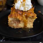 Slice of pumpkin bread pudding garnished with whipped cream and salted caramel sauce on a dark surface.