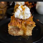 Slice of pumpkin bread pudding garnished with whipped cream and salted caramel sauce on a dark surface.