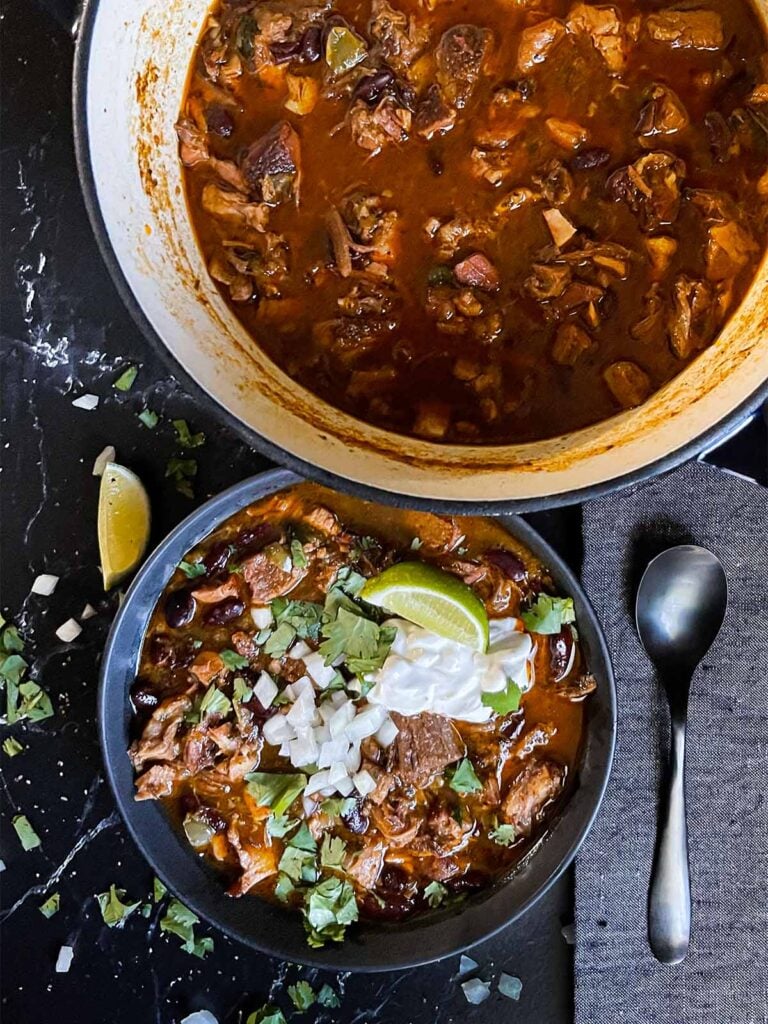 Smoked pork chili garnished with cilantro, onion, lime, and onion in a dark bowl.