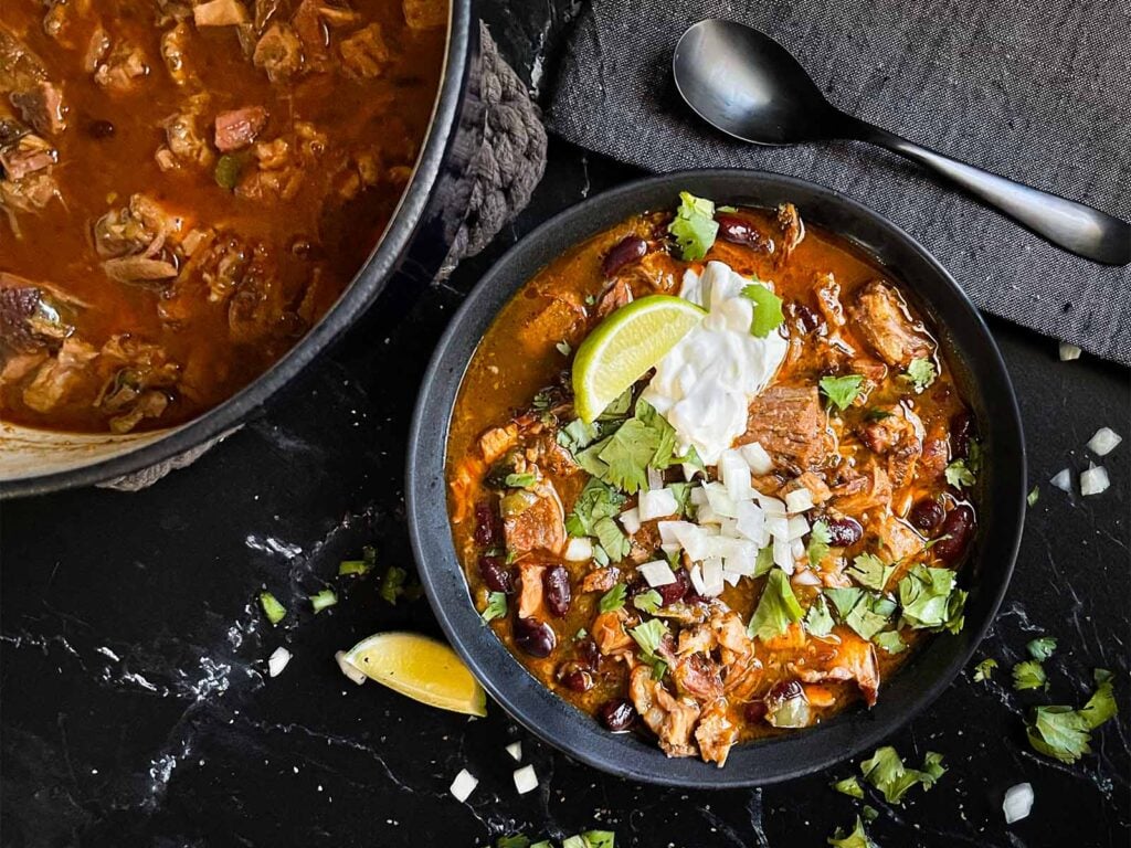 Smoked pork chili garnished with cilantro, onion, lime, sour cream, and onion in a dark bowl.