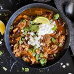 Smoked pork chili garnished with cilantro, onion, lime, sour cream, and onion in a dark bowl.