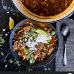 Smoked pork chili garnished with cilantro, onion, lime, sour cream, and onion in a dark bowl.