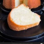 A slice of pound cake on a black plate with the whole pound cake in the background.