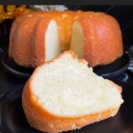 A slice of pound cake on a black plate with the whole pound cake in the background.