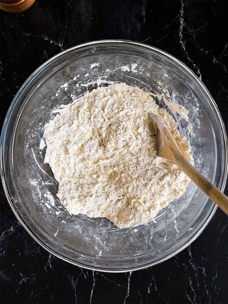 The grated butter tossed with the flour mixture for sweet potato biscuits in a glass bowl on a dark surface.