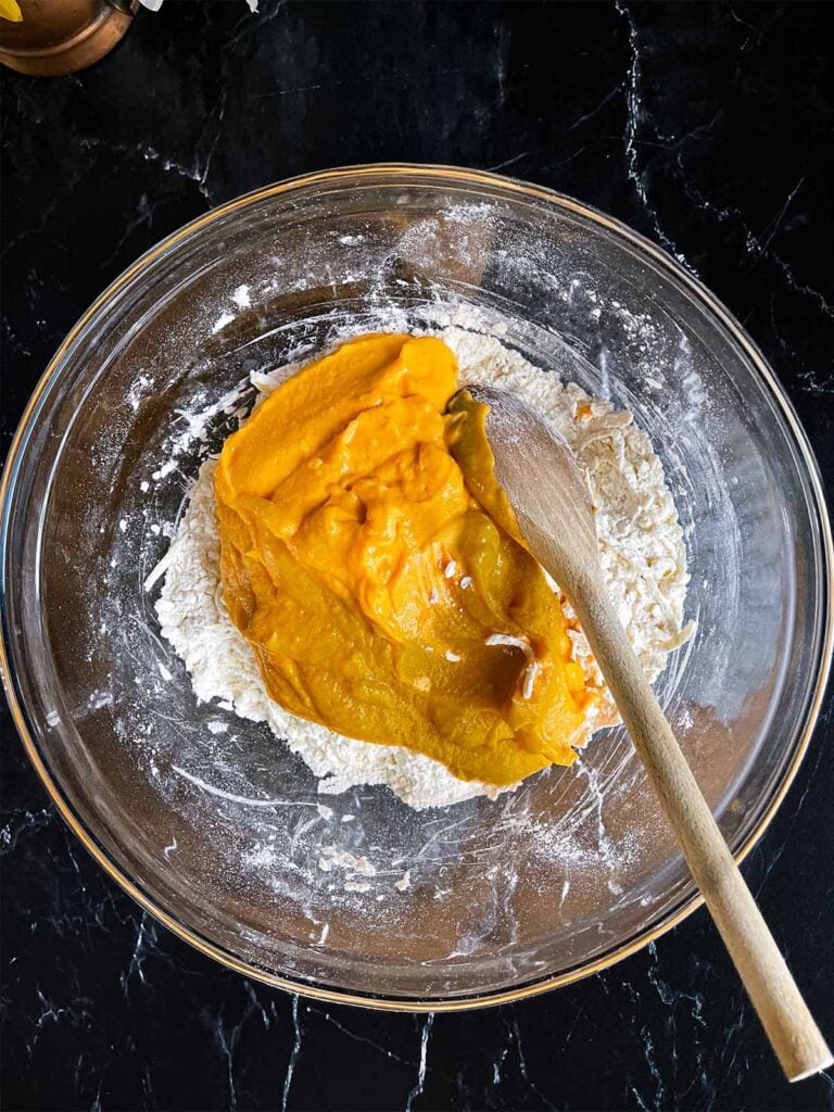 Sweet potato mixture added to the flour and butter mixture for sweet potato biscuits in a glass mixing bowl on a dark surface.