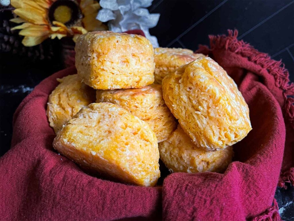 Baked sweet potato biscuits stacked in a bread basket, lined with a cloth napkin on a dark surface.