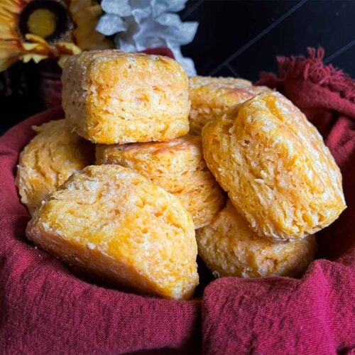 Baked sweet potato biscuits stacked in a bread basket, lined with a cloth napkin on a dark surface.