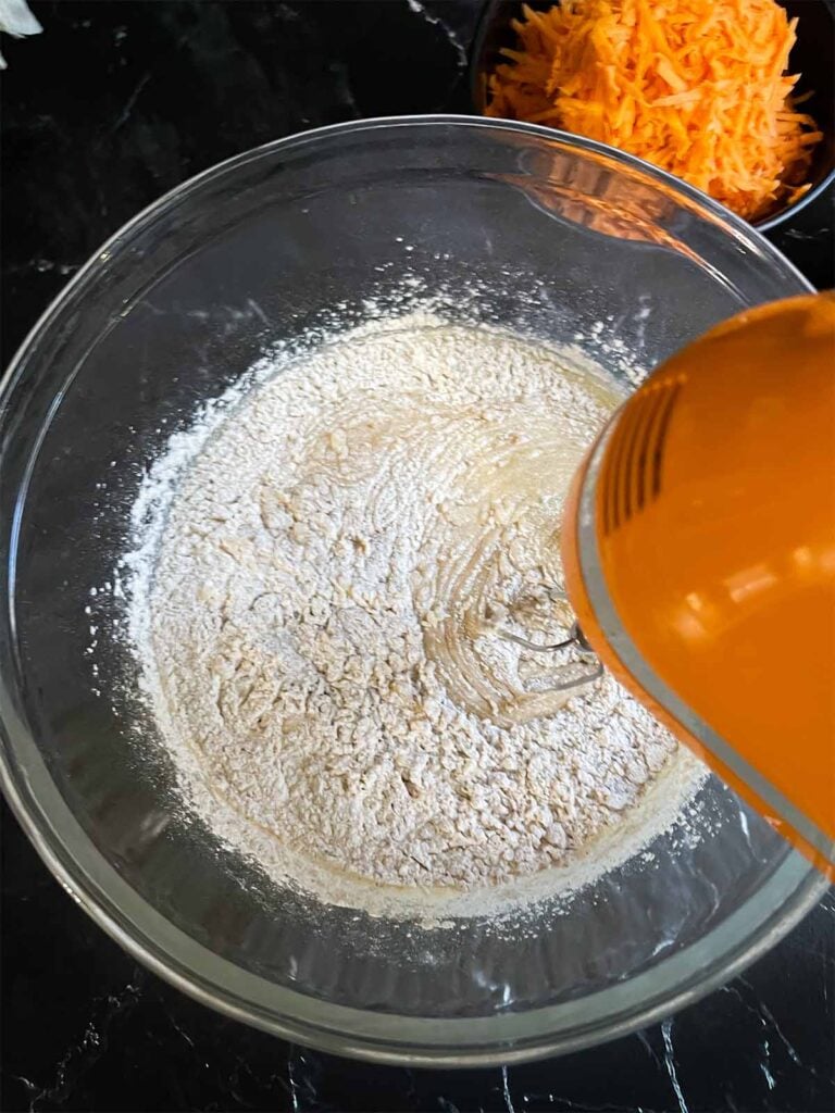 Dry ingredients being added to the wet ingredients for sweet potato cake in a glass mixing bowl on a dark surface.