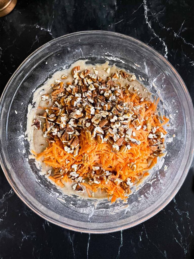 Shredded sweet potato and toasted pecans added to the sweet potato cake mixture in a glass mixing bowl.