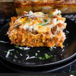 A serving of baked spaghetti on a dark plate with the casserole in the background.