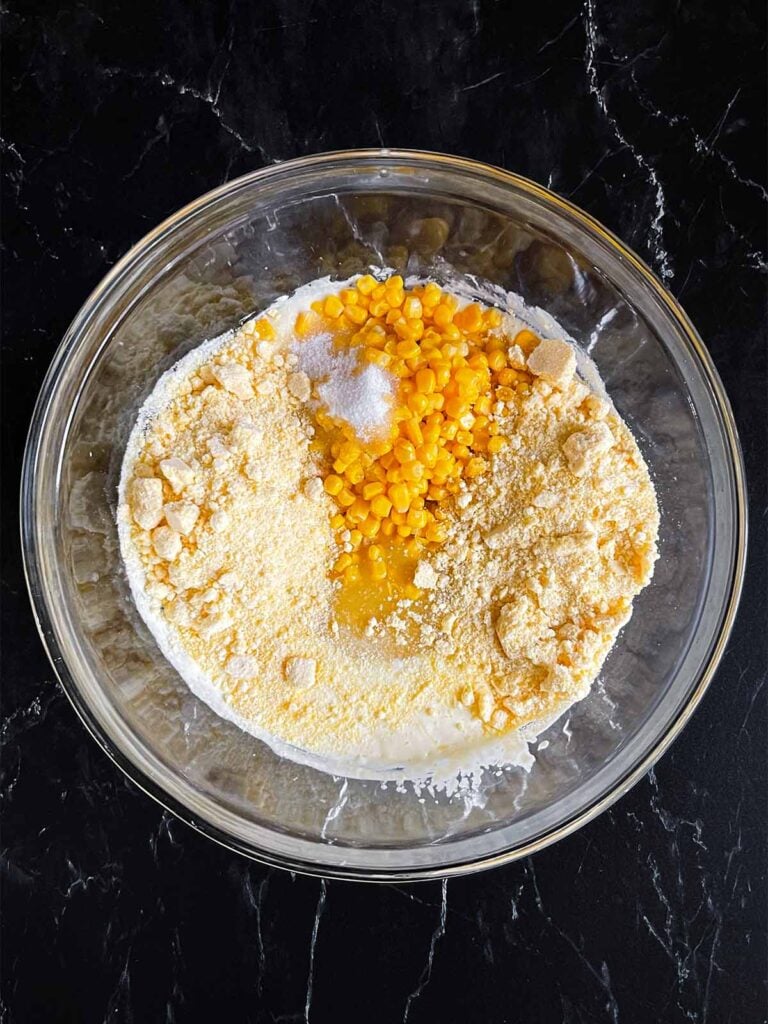 Corn and dry ingredients added to the wet ingredients in a glass bowl.
