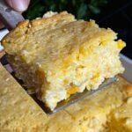 A serving of creamy corn casserole lifted over the baking pan on a dark surface.