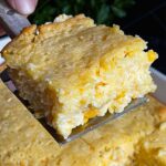 A serving of creamy corn casserole lifted over the baking pan on a dark surface.