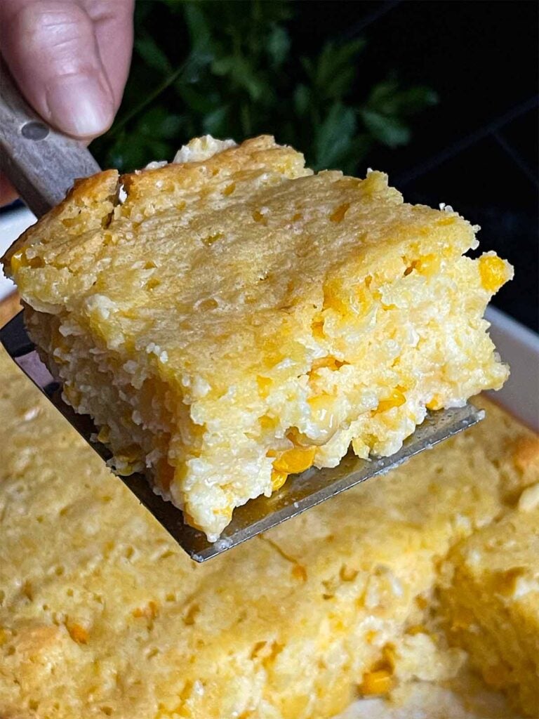 A serving of creamy corn casserole lifted over the baking pan on a dark surface.