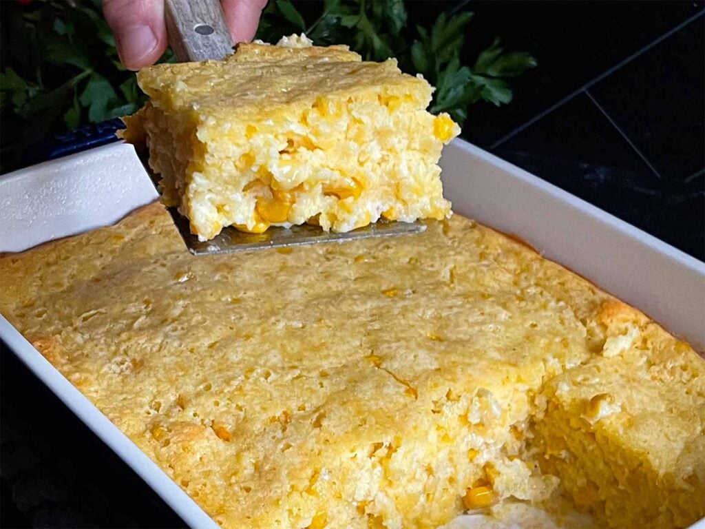 A serving of creamy corn casserole lifted over the baking pan on a dark surface.