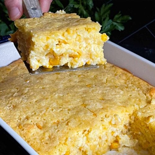 A serving of creamy corn casserole lifted over the baking pan on a dark surface.